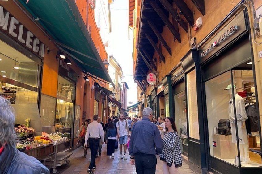 Una calle estrecha flanqueada por tiendas, con peatones caminando y diversos productos exhibidos. 1