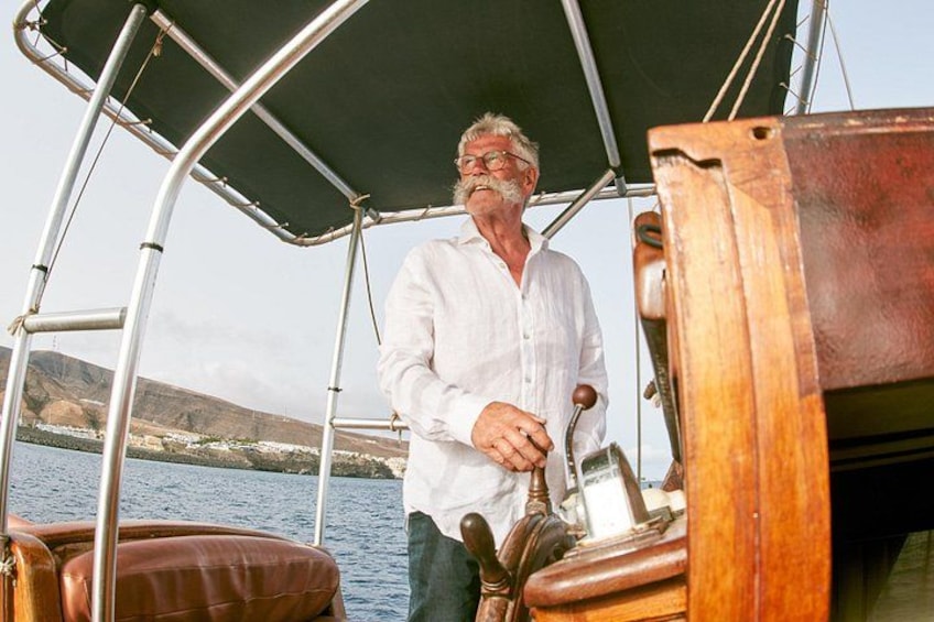 A man on a boat with a steering wheel, a canopy, and a rocky coastline in the background. 5