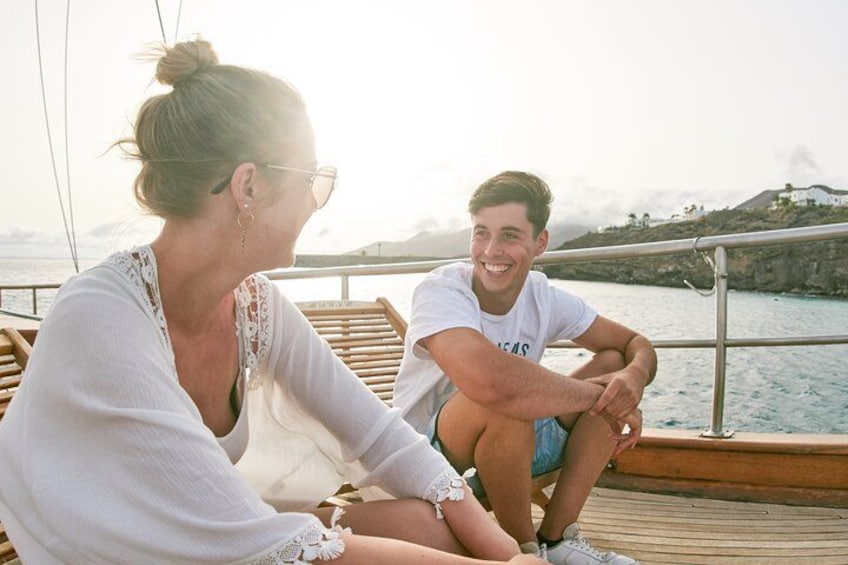 Two people sitting on a wooden deck by a body of water with a rocky coastline in the background. 3
