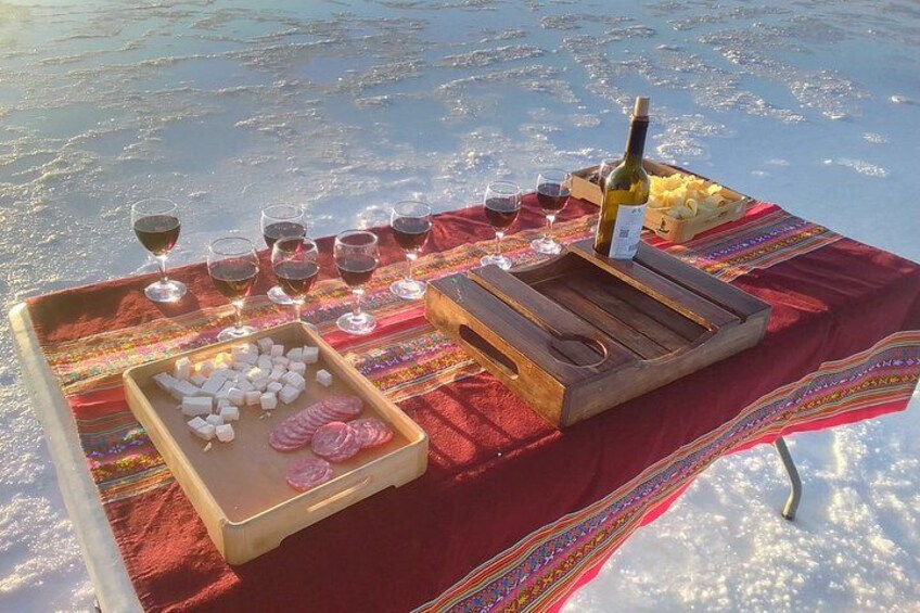 A picnic setup on a snowy surface with a tablecloth, wine glasses, a bottle, and snacks. 3