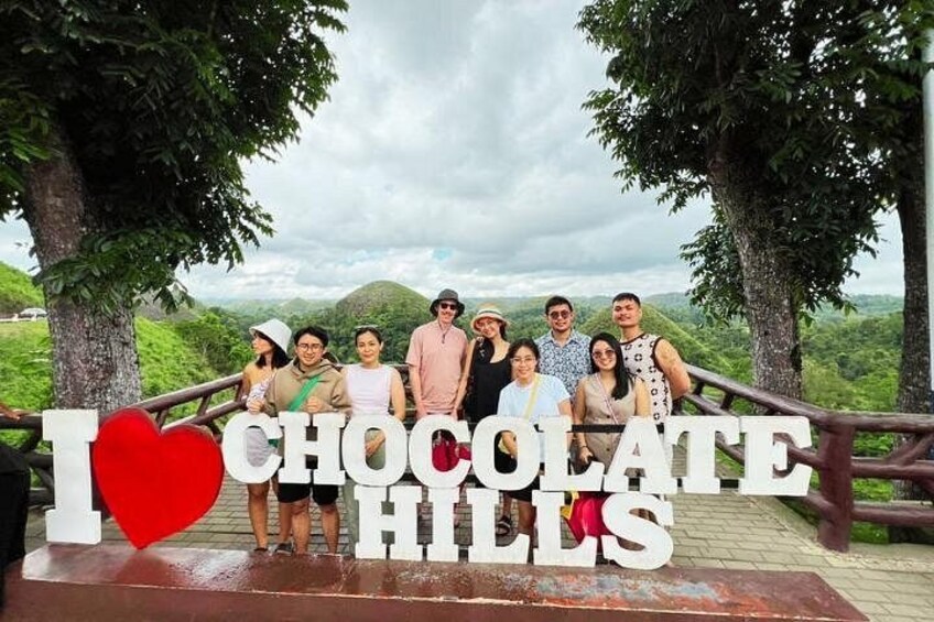 Eine Gruppe von Menschen steht in einer natürlichen Umgebung, mit einem Schild mit der Aufschrift 'I ♥ CHOCOLATE HILLS'. 1