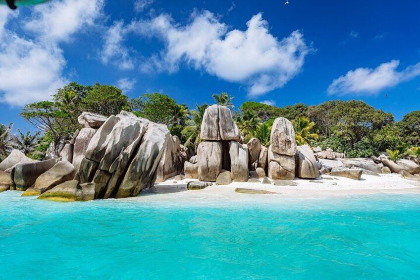 Ein Strand mit großen weißen Felsen, klarem türkisblauem Wasser und grünen Palmen unter blauem Himmel. 1