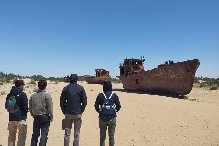 Four people standing on a sandy beach, looking at a large rusted shipwreck. 3