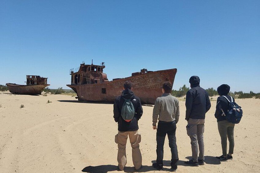 Four people standing on a sandy beach, looking at a large rusted shipwreck. 2
