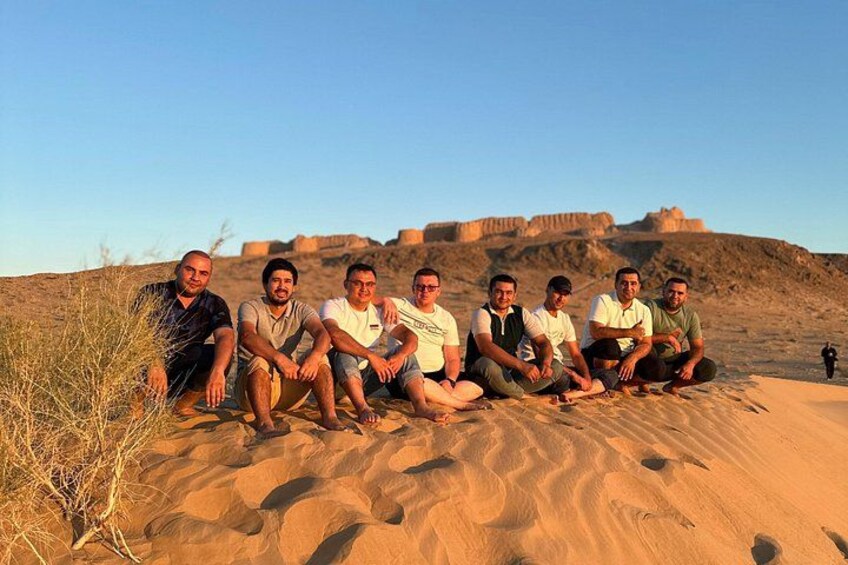 A group of people sitting on a sandy dune with a fort in the background. 1