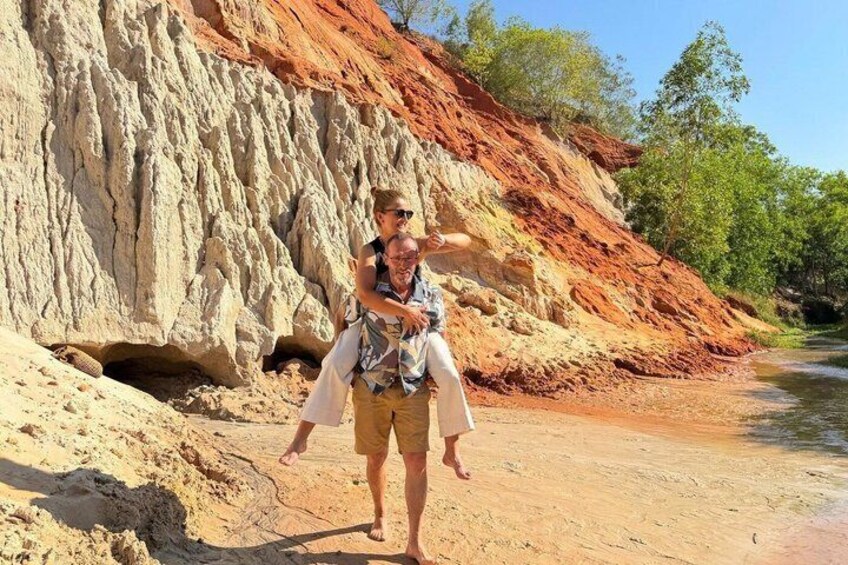 A person carrying another on their back, walking on a sandy path with red earth formations in the background. 2