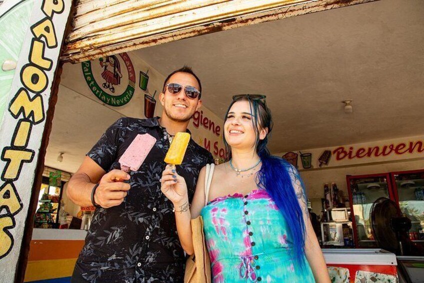 Two people holding ice cream bars in front of a shop with a 'Bienvenidos' sign. 3