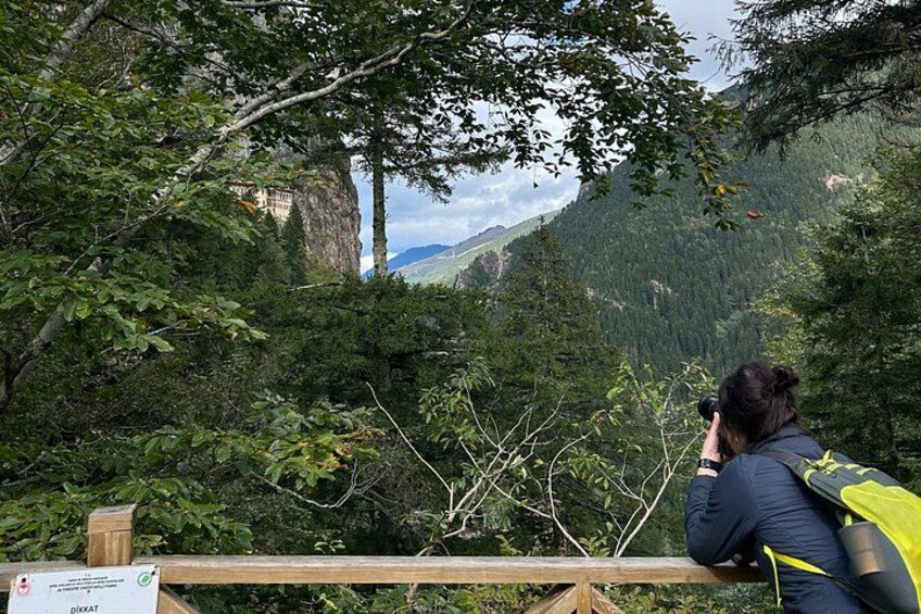 Una persona fotografiando un paisaje montañoso con bosque denso y un edificio al fondo. 1