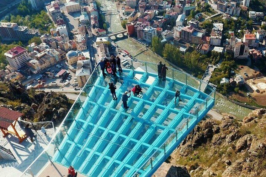 A blue glass bridge with people walking on it, overlooking a cityscape with buildings and a river. 3