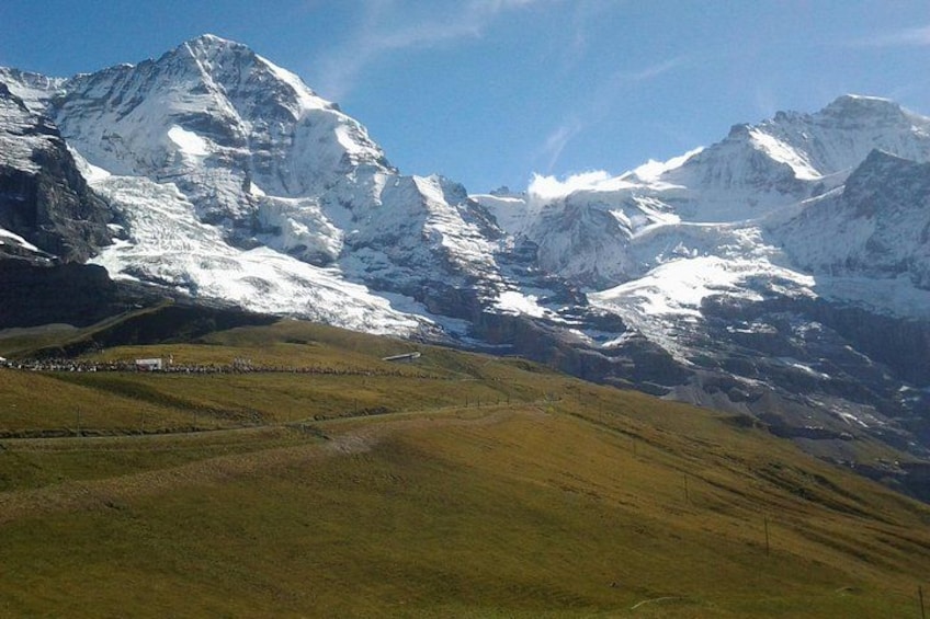 A mountainous landscape with snow-capped peaks, a valley with green grass, and a small settlement. 2