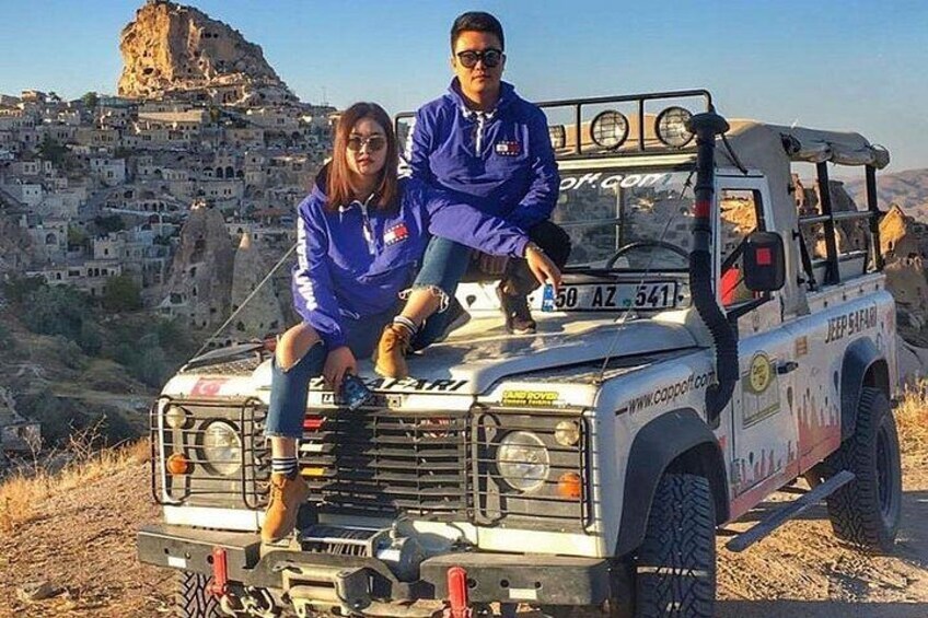 A Jeep Safari with two people sitting on the hood, a mountainous town in the background, and a clear sky. 5
