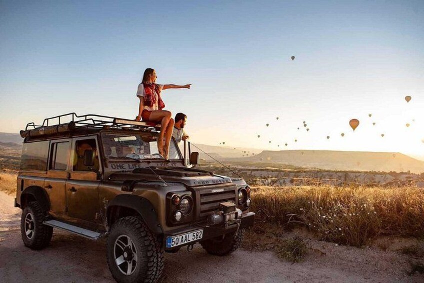 A vehicle with a license plate, a person standing on the roof, and hot air balloons in the background. 1