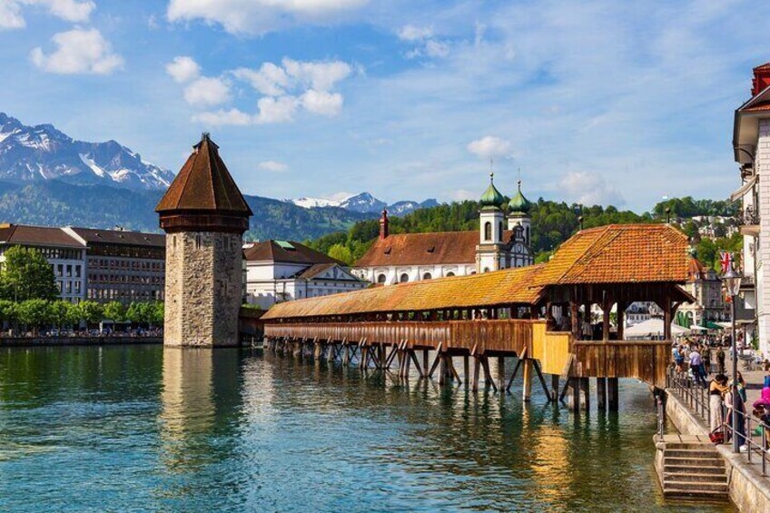 Un pueblo a orillas del río con un puente de madera, una torre y edificios de techos verdes. 1