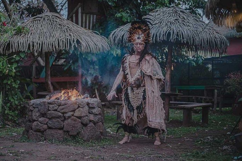 A person in traditional attire standing near a fire pit with thatched umbrellas in a garden setting. 2