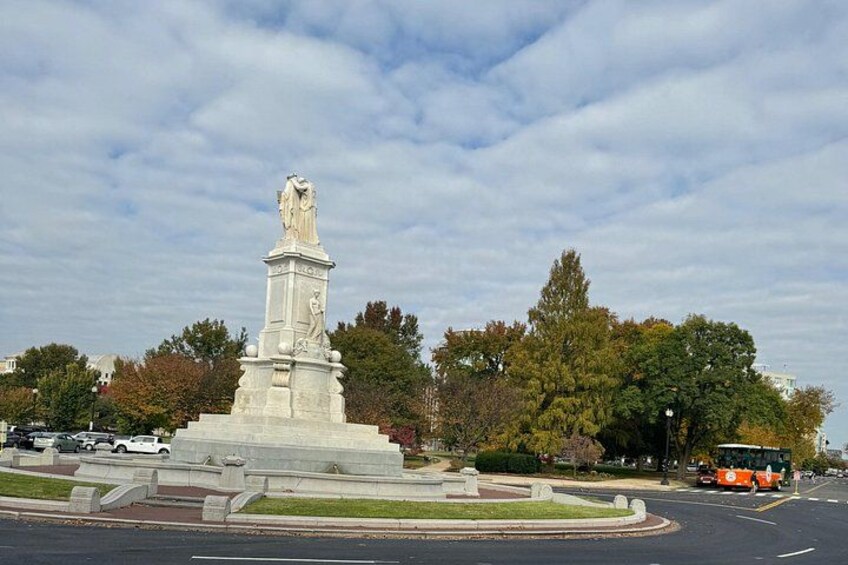 Un monumento blanco con una estatua, rodeado de un área ajardinada con árboles y una carretera con vehículos. 5
