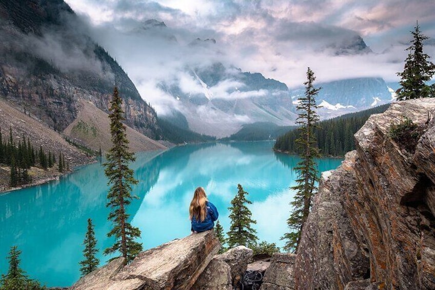 A person sitting on a rock overlooking a turquoise lake surrounded by mountains and trees. 1