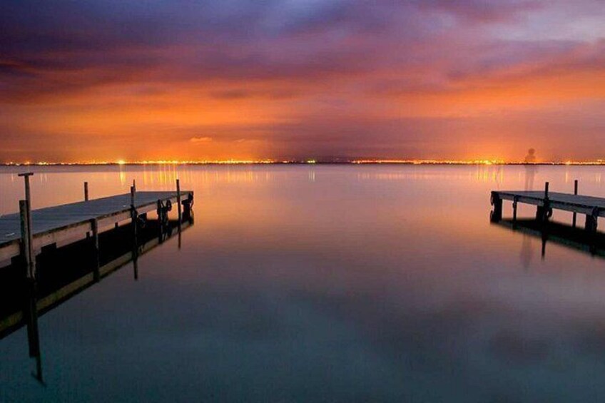 A calm waterfront at dusk with a wooden dock extending into the water, a clear sky, and distant city lights. 2