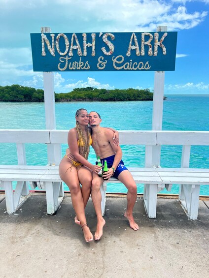 Two people sitting on a bench by a sign that reads "NOAH'S ARK Turks & Caicos." 3