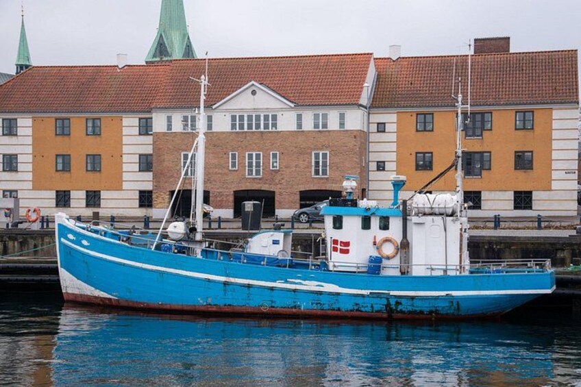 En blå fiskebåd ligger fortøjet i en havn med bygninger i baggrunden. 4