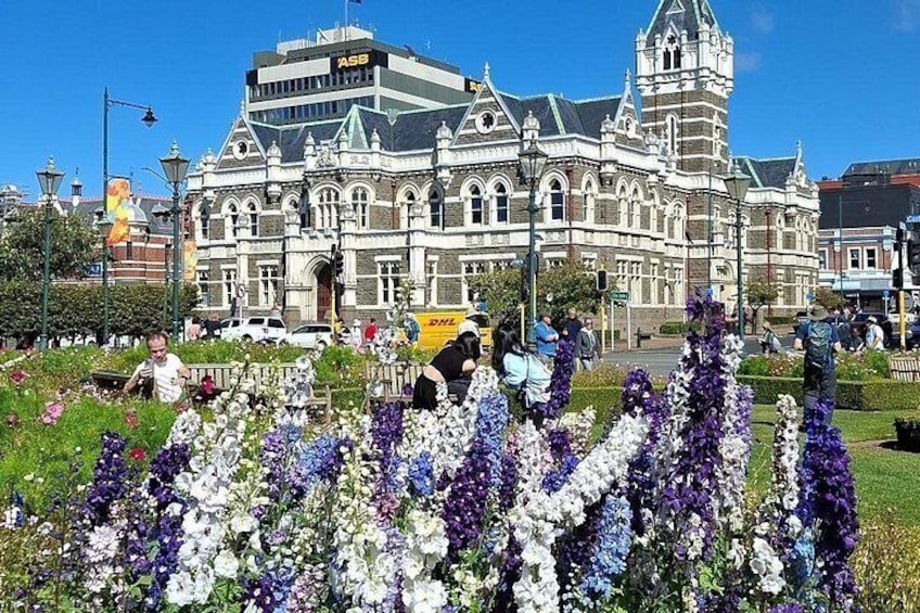 A historic building with ornate architecture, a flower garden in the foreground, and a DHL delivery truck in the background. 3