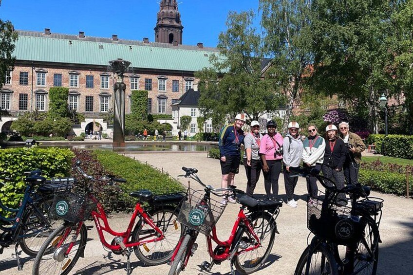En grupp människor som står framför cyklar, med en historisk byggnad och en fontän i bakgrunden. 4