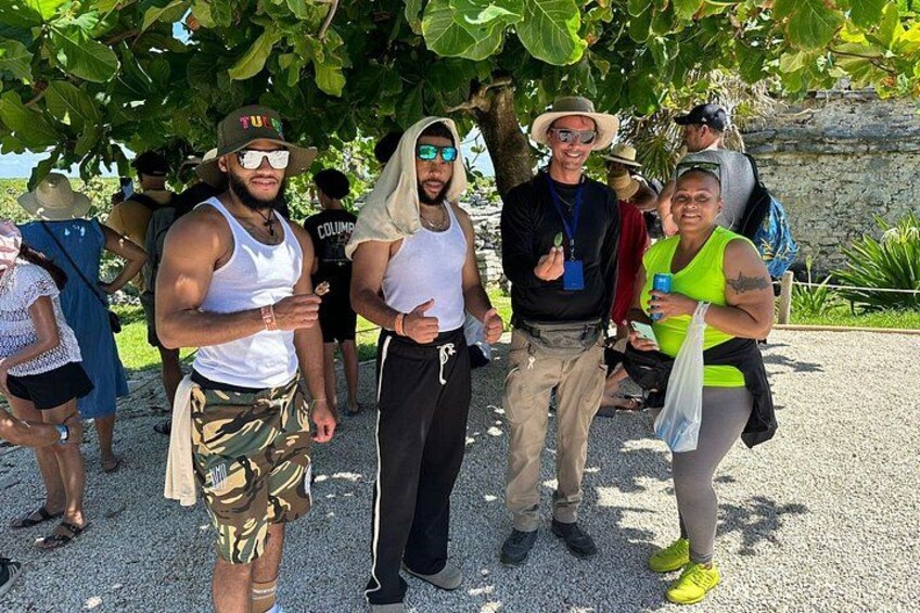 A group of people standing outdoors, some wearing hats and sunglasses, under a tree with a stone wall in the background. 4
