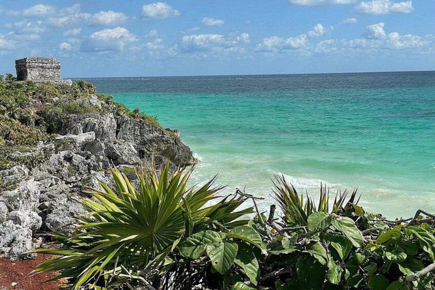 A coastal landscape with clear turquoise waters, a rocky cliff, and a small ancient structure. 2