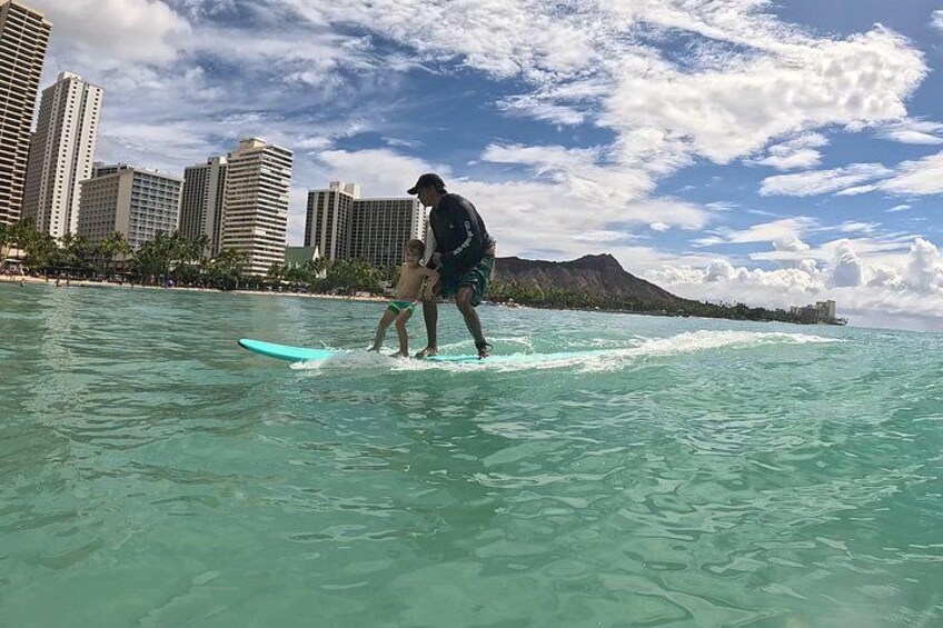 A person surfing with a child in the ocean, with a cityscape and mountains in the background. 4