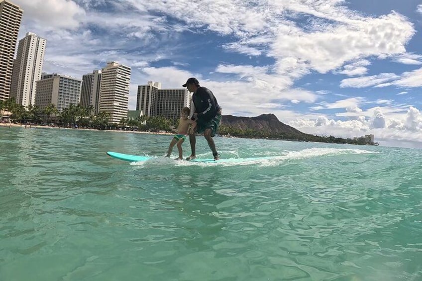 Eine Person surft mit einem Kind im Meer, im Hintergrund eine Stadtansicht und ein Berg. 3