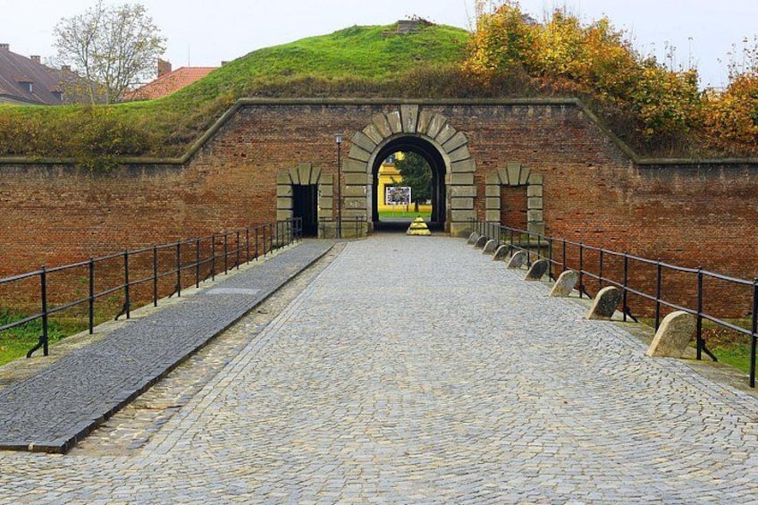 A cobblestone pathway leading to an arched entrance with a brick wall and a grassy hill in the background. 3