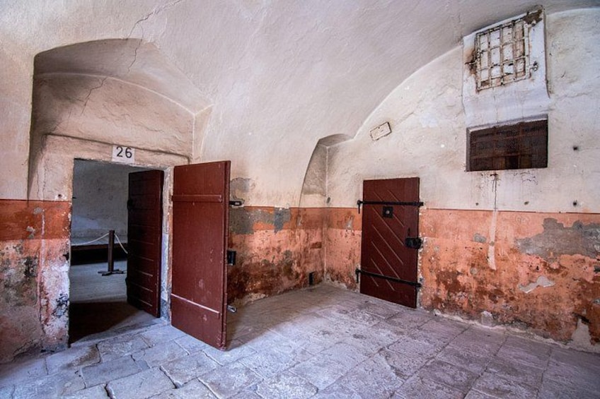 An arched corridor with a number 26 sign, a wooden door, and a barred window. 5