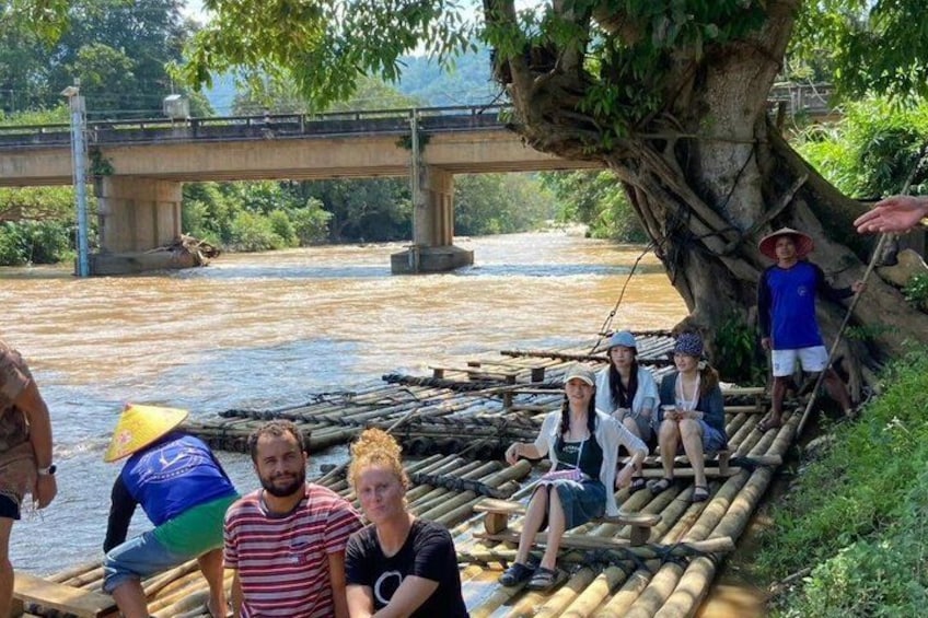 People on a bamboo raft on a river, a bridge in the background, and a tree with roots overhanging the water. 3