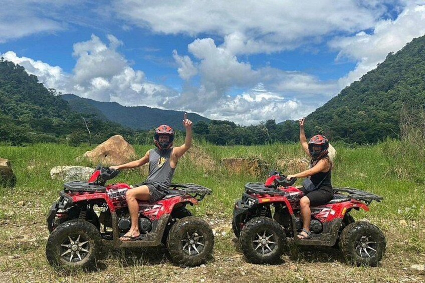 Two people on red ATVs in a grassy field with mountains in the background. 1