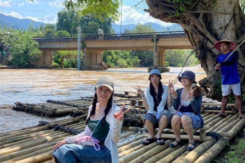 People sitting on bamboo rafts by a river with a bridge in the background. 4