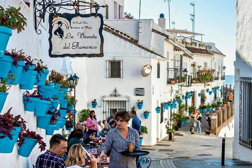 A narrow street with white buildings, blue planters, and a sign reading 'La Boveda del Flamenco'. 1