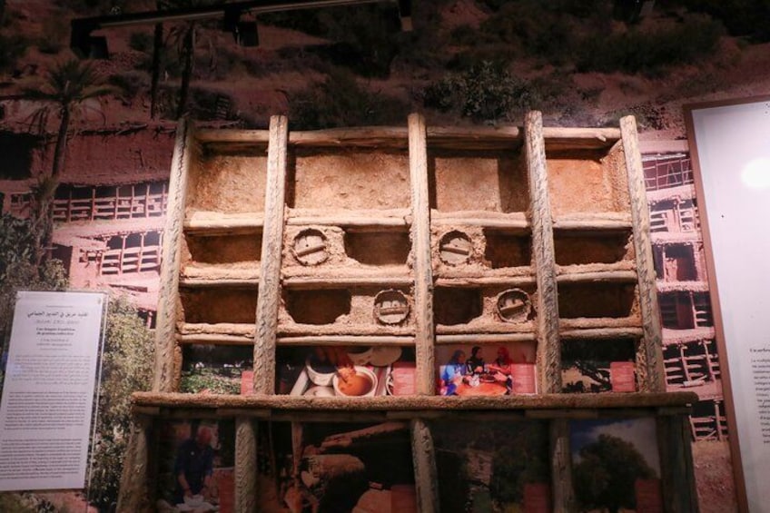 Una vitrina de madera con estantes, algunos objetos en el estante inferior, y una pared con texto e imágenes al fondo. 3