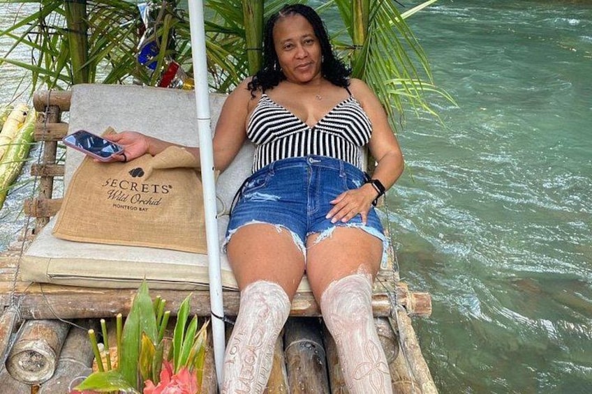 A person relaxing on a wooden platform by water, wearing a striped top and denim shorts, with a bag labeled 'SECRETS Wild Orchid' and a phone. 2