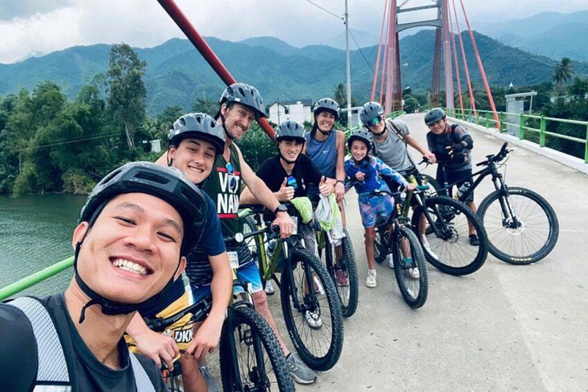 Un groupe de personnes se trouve sur un pont, avec des vélos, une rivière et des montagnes en arrière-plan. 3