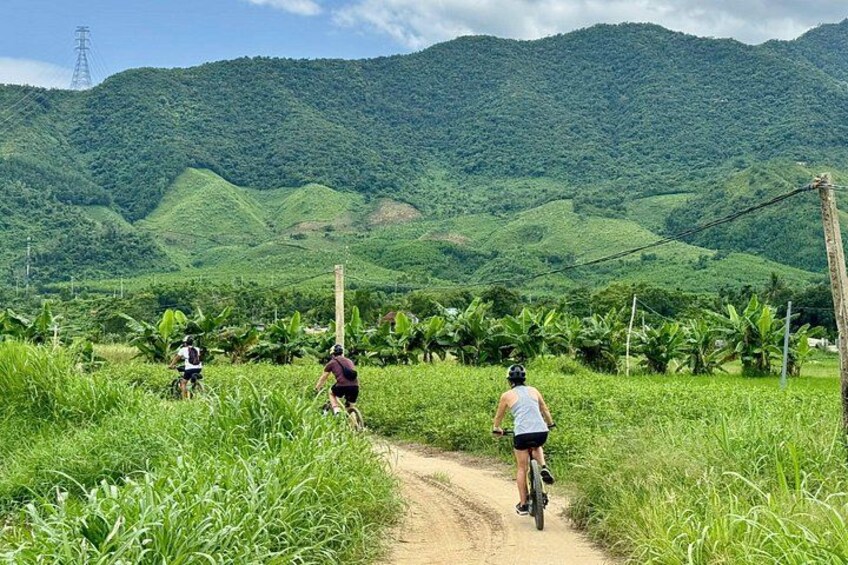 Un chemin de terre bordé de cyclistes, de collines verdoyantes et de plants de bananiers. 1