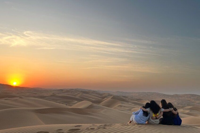 A sunset over sand dunes with three people sitting and embracing. 1