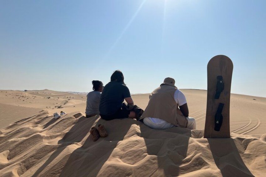 Three people sitting on sand dunes with a snowboard leaning against them. 4