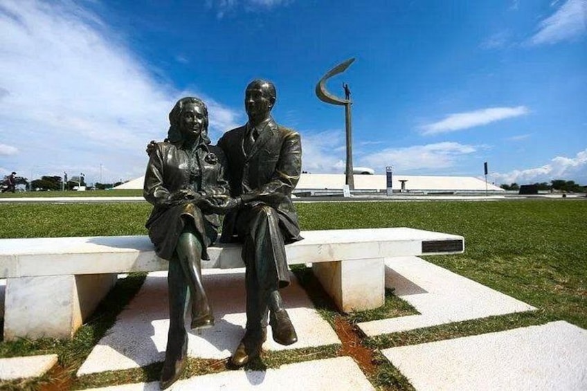 Eine Statue von einem Mann und einer Frau, die auf einer Bank in einem Park sitzen, mit einer modernen Skulptur im Hintergrund. 3
