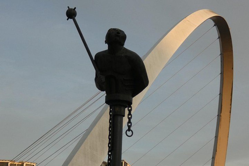 Una estatua de una figura sosteniendo un báculo, montada sobre un poste, con una gran estructura en forma de arco al fondo. 3