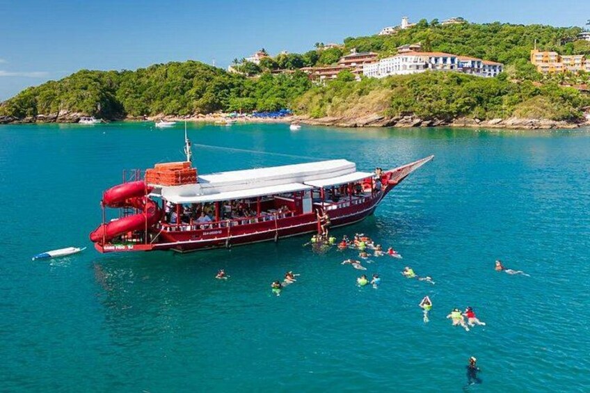 Un bateau rouge transportant des passagers, entouré d’une eau d’un bleu limpide et d’un paysage vallonné avec des bâtiments en arrière-plan. 2