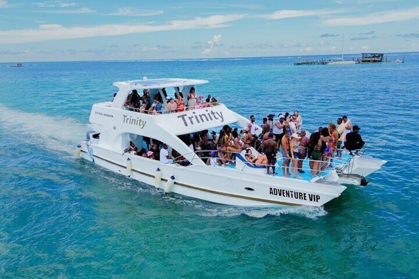 En hvid båd, der hedder 'Trinity', med teksten 'Adventure VIP' på siden, fyldt med mennesker, der nyder vandet. 1