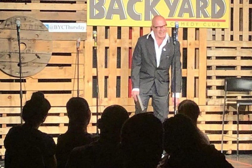 Un homme en costume, debout sur une scène, avec une enseigne indiquant « BACKYARD COMEDY CLUB » en arrière-plan. 5