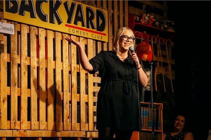 A person performing on stage at a comedy club with a wooden backdrop and a microphone stand. 2