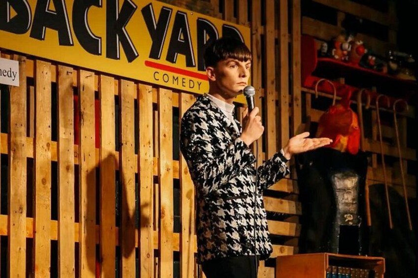 Un artiste qui fait du stand-up dans un lieu où l’on peut voir, en arrière-plan, un décor en bois et une pancarte sur laquelle est écrit : « COMÉDIE EN PLEIN AIR ». 3