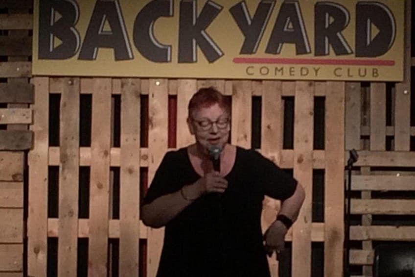 Une personne tenant un microphone devant une enseigne sur laquelle on peut lire « BACKYARD COMEDY CLUB ». 3