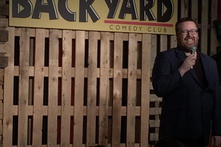 Un homme tenant un microphone devant une clôture en bois sur laquelle est affiché un panneau indiquant « BACKYARD COMEDY CLUB ». 5